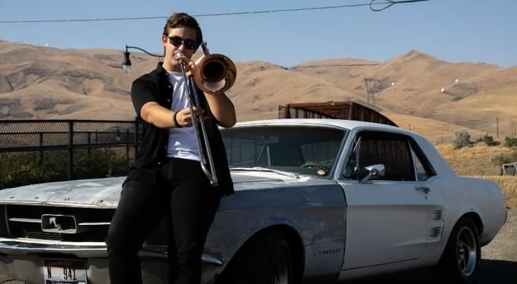 Lane Riggs plays his instrument while sitting by his car.